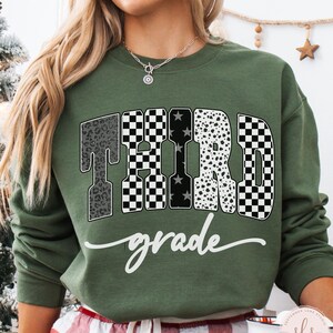 May include: An olive green sweatshirt with the words "THIRD grade" in a mix of patterns, including leopard print, checkered, and polka dots. The word "grade" is in a cursive font. The sweatshirt is a casual style.