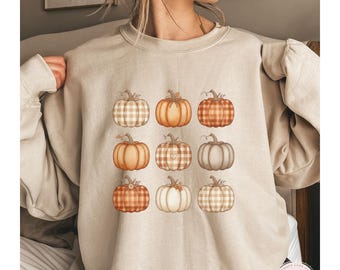 May include: A sand-coloured sweatshirt displaying a grid of nine watercolour pumpkins in shades of orange, brown, and cream. The pumpkins feature diverse patterns, including plaid and solid colours. The word "SAND" is printed in capital letters at the bottom left.