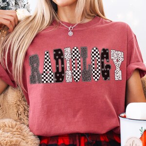 K&ouml;nnte beinhalten: Karmesinrotes T-Shirt mit dem Wort "RADIOLOGY" in einem auff&auml;lligen, gemusterten Design. Die Buchstaben weisen einen Mix aus Leopardenmuster, Sternen und Schachbrettmustern auf. Das Shirt ist ein bequemer, l&auml;ssiger Stil.