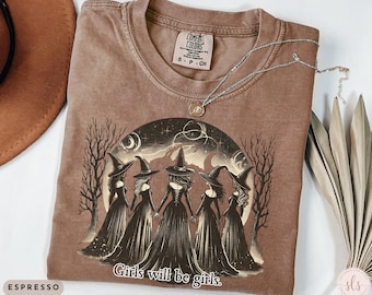 May include: Espresso-colored t-shirt featuring a graphic of five witches holding hands under a night sky with the text "Girls will be girls." The shirt has a crew neck and short sleeves. A silver necklace with a pendant is draped across the shirt.
