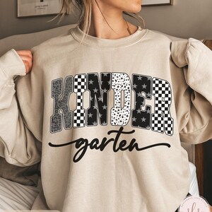 May include: Sand-colored sweatshirt with the word "KINDER" in a bold, patterned design, including leopard print, stars, and checkered patterns. The word "garten" is written in a cursive font below the main text.