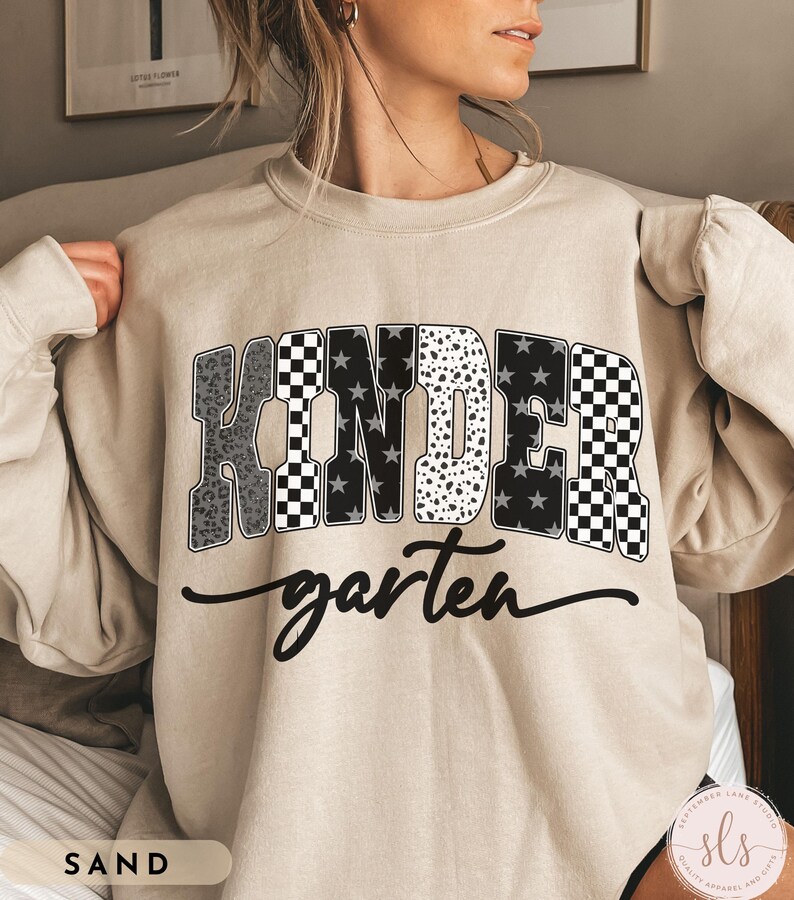 May include: Sand-colored sweatshirt with the word "KINDER" in a bold, patterned design, including leopard print, stars, and checkered patterns. The word "garten" is written in a cursive font below the main text.
