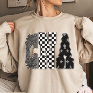 May include: A sand-colored sweatshirt featuring the letters "CNA" in a bold, eye-catching design. The "C" has a leopard print pattern, the "N" is checkered, and the "A" is filled with stars. The word "SAND" is printed at the bottom.