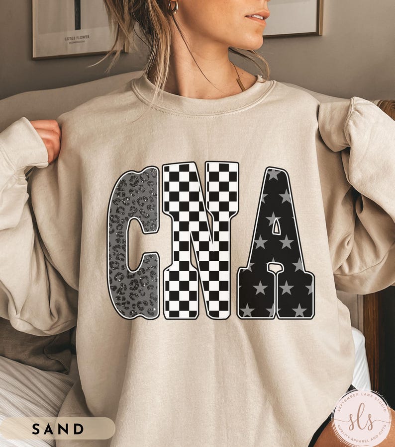 May include: A sand-colored sweatshirt featuring the letters "CNA" in a bold, eye-catching design. The "C" has a leopard print pattern, the "N" is checkered, and the "A" is filled with stars. The word "SAND" is printed at the bottom.