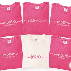 May include: A collection of pink t-shirts with white script text, including "Mother of the Bride," "Bridesmaid," "Mother of the Groom," "Matron of Honor," "Bride," and "Maid of Honor." One shirt is cream-colored. The shirts are arranged in a group.