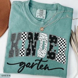 May include: Light green t-shirt with the word "KINDERgarten" in a bold, multi-patterned design. The shirt features a crew neck and short sleeves. A gold necklace with a pendant is draped across the shirt.