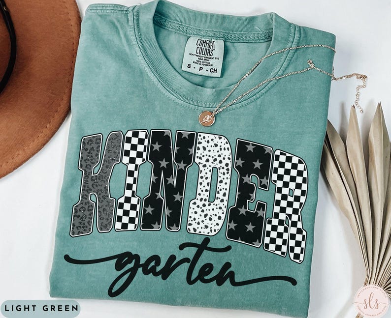 May include: Light green t-shirt with the word "KINDERgarten" in a bold, multi-patterned design. The shirt features a crew neck and short sleeves. A gold necklace with a pendant is draped across the shirt.