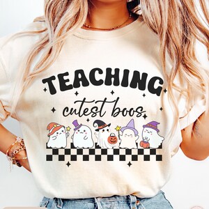 May include: Ivory-colored t-shirt with the text "TEACHING cutest boos" in black. Below the text are five cartoon ghosts wearing witch hats and holding pumpkins. A black and white checkered pattern is at the bottom.