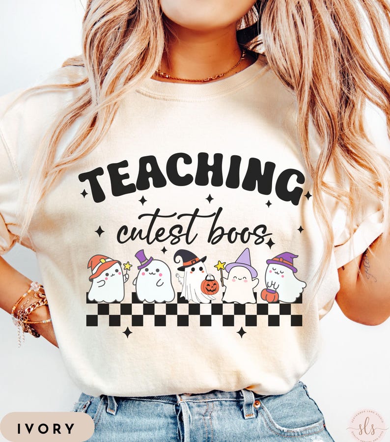 May include: Ivory-colored t-shirt with the text "TEACHING cutest boos" in black. Below the text are five cartoon ghosts wearing witch hats and holding pumpkins. A black and white checkered pattern is at the bottom.