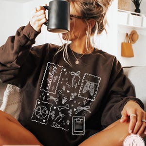 Puede incluir: Sudadera de color chocolate oscuro con ilustraciones gr&aacute;ficas blancas de im&aacute;genes m&eacute;dicas, incluyendo un esqueleto, huesos y un s&iacute;mbolo de radiaci&oacute;n. La palabra "radiology" est&aacute; escrita en una fuente decorativa. La sudadera tiene mangas largas.