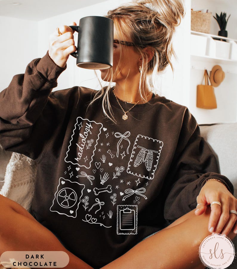 Puede incluir: Sudadera de color chocolate oscuro con ilustraciones gr&aacute;ficas blancas de im&aacute;genes m&eacute;dicas, incluyendo un esqueleto, huesos y un s&iacute;mbolo de radiaci&oacute;n. La palabra "radiology" est&aacute; escrita en una fuente decorativa. La sudadera tiene mangas largas.