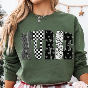 May include: A military green sweatshirt with the word "NURSE" in a bold, patterned design. The letters feature a mix of leopard print, checkered, star, and dalmatian patterns. The sweatshirt is a crew neck style.