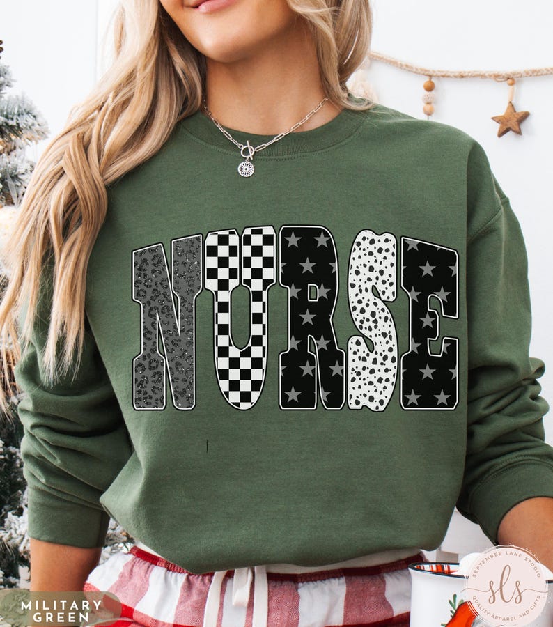 May include: A military green sweatshirt with the word "NURSE" in a bold, patterned design. The letters feature a mix of leopard print, checkered, star, and dalmatian patterns. The sweatshirt is a crew neck style.