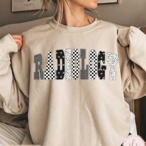 K&ouml;nnte beinhalten: Sandfarbenes Sweatshirt mit dem Wort "RADIOLOGY" in einem auff&auml;lligen, gemusterten Design. Die Buchstaben weisen eine Mischung aus schwarz-wei&szlig; karierten, Leopardenmuster- und Sternmustern auf. Das Sweatshirt hat eine neutrale Farbe.