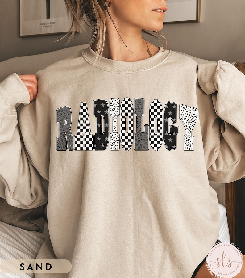 K&ouml;nnte beinhalten: Sandfarbenes Sweatshirt mit dem Wort "RADIOLOGY" in einem auff&auml;lligen, gemusterten Design. Die Buchstaben weisen eine Mischung aus schwarz-wei&szlig; karierten, Leopardenmuster- und Sternmustern auf. Das Sweatshirt hat eine neutrale Farbe.