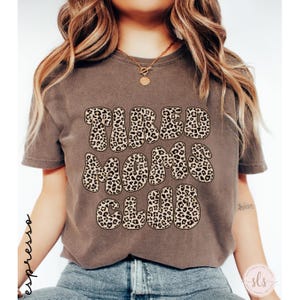 K&ouml;nnte beinhalten: Ein braunes T-Shirt mit dem Text "TIRED MOMS CLUB" im Leopardenmuster. Das Shirt hat kurze &Auml;rmel und einen Rundhalsausschnitt. Das Wort "espresso" steht vertikal auf der linken Seite.