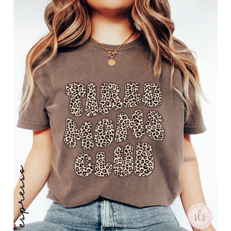 K&ouml;nnte beinhalten: Ein braunes T-Shirt mit dem Text "TIRED MOMS CLUB" im Leopardenmuster. Das Shirt hat kurze &Auml;rmel und einen Rundhalsausschnitt. Das Wort "espresso" steht vertikal auf der linken Seite.