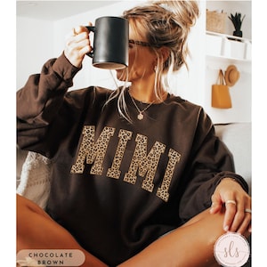 May include: A chocolate brown sweatshirt featuring the word "MIMI" in a leopard print pattern. The crew neck sweatshirt has long sleeves. The image also shows a black mug and a small logo in the bottom right corner.