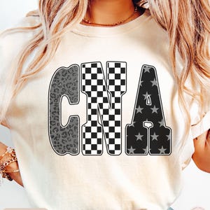 May include: Ivory-colored t-shirt with the letters "CNA" in a bold, outlined font. The "C" has a gray leopard print, the "N" is a black and white checkerboard pattern, and the "A" features gray stars on a black background. The word "IVORY" is at the bottom.