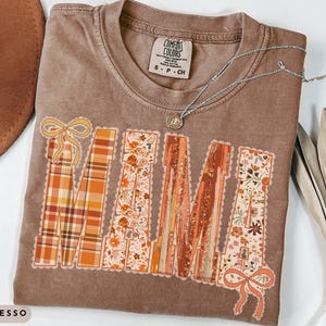 May include: Espresso-colored Comfort Colors t-shirt with the word "MAMA" in a decorative design. The letters feature plaid, floral, and striped patterns in shades of orange, brown, and cream. A silver necklace with a pendant rests on the shirt.
