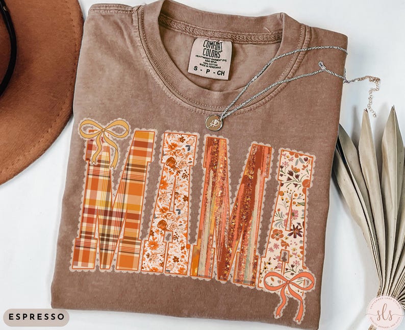 May include: Espresso-colored Comfort Colors t-shirt with the word "MAMA" in a decorative design. The letters feature plaid, floral, and striped patterns in shades of orange, brown, and cream. A silver necklace with a pendant rests on the shirt.