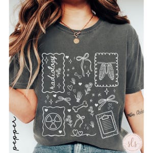 Puede incluir: Camiseta gris oscuro con un dise&ntilde;o gr&aacute;fico blanco que presenta la palabra "radiolog&iacute;a", huesos, lazos, corazones y una caja tor&aacute;cica. La camiseta tiene un corte holgado y mangas cortas.