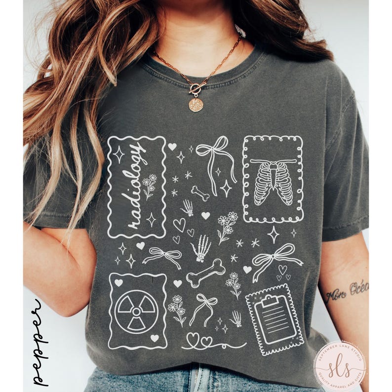 Puede incluir: Camiseta gris oscuro con un dise&ntilde;o gr&aacute;fico blanco que presenta la palabra "radiolog&iacute;a", huesos, lazos, corazones y una caja tor&aacute;cica. La camiseta tiene un corte holgado y mangas cortas.