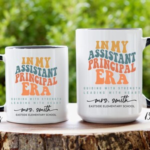 May include: Two white ceramic coffee mugs with black handles. The mugs have a colorful retro design with the text "In My Assistant Principal Era" and "Guiding with Strength Leading with Heart". The text also includes the name "mrs. smith" and "Eastside Elementary School". The mugs are 11 oz and 15 oz.