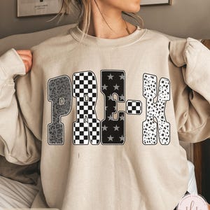May include: Sand-colored sweatshirt with the word "PRE-K" in a bold, patterned design. The letters feature leopard print, a black and white checkerboard, stars, and dalmatian spots. The sweatshirt has long sleeves.