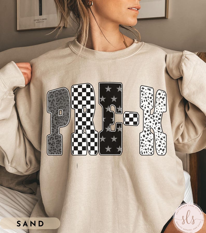 May include: Sand-colored sweatshirt with the word "PRE-K" in a bold, patterned design. The letters feature leopard print, a black and white checkerboard, stars, and dalmatian spots. The sweatshirt has long sleeves.