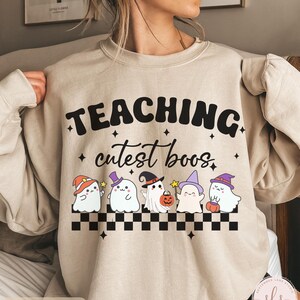 May include: Sand-colored sweatshirt with the text "TEACHING cutest boos" in black. Below the text are five cartoon ghosts wearing Halloween hats and holding a pumpkin and wands. A black and white checkered pattern is at the bottom.