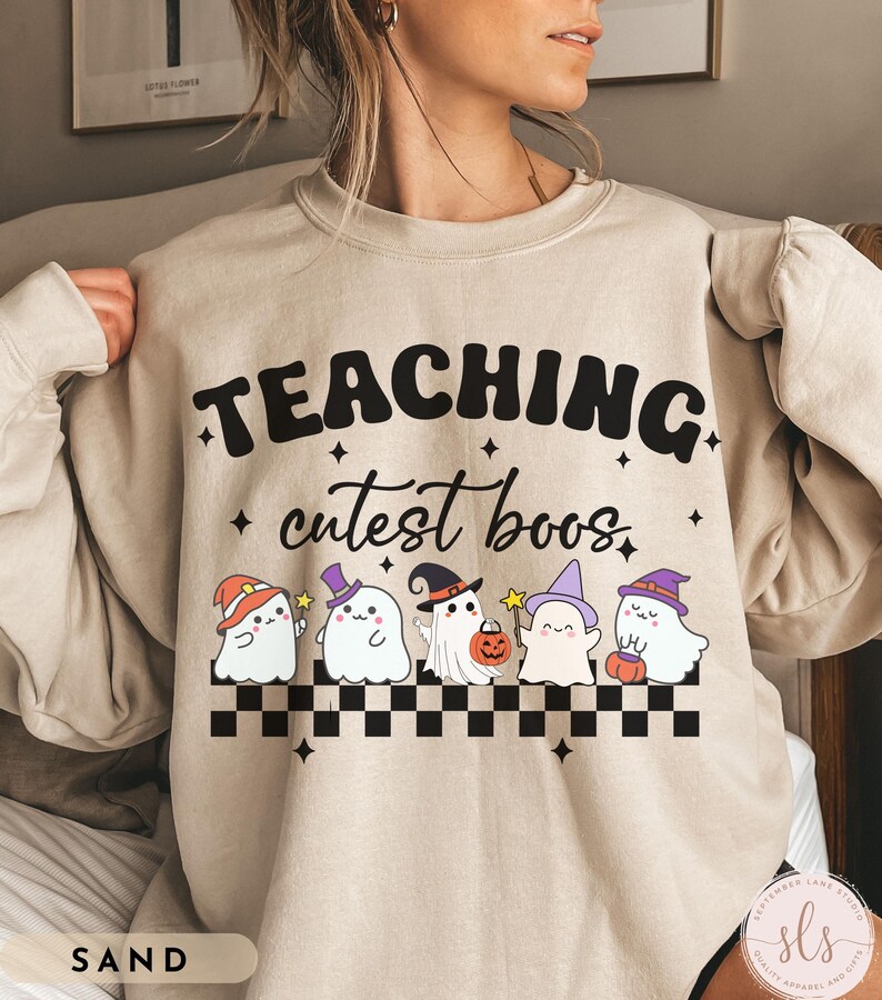 May include: Sand-colored sweatshirt with the text "TEACHING cutest boos" in black. Below the text are five cartoon ghosts wearing Halloween hats and holding a pumpkin and wands. A black and white checkered pattern is at the bottom.