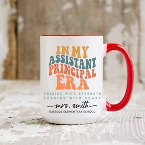 May include: White ceramic coffee mug with a red rim and handle. The mug has a colorful graphic that says "In My Assistant Principal Era" with the text "Guiding with Strength Leading with Heart" and "mrs. smith Eastside Elementary School".