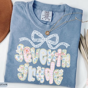 May include: Blue jean Comfort Colors t-shirt with a floral and gingham "Seventh Grade" design. The shirt features a white bow graphic and a gold necklace. The shirt is size small.