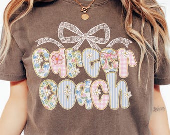 May include: A brown t-shirt featuring the words "Career Coach" in a colourful design with floral and gingham patterns. The text is outlined in gold and topped with a white lace bow.