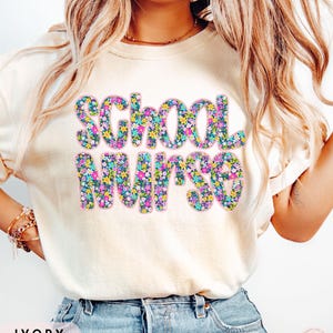 May include: Ivory-colored t-shirt with the words "School Nurse" in a floral design. The text is filled with colorful flowers in shades of blue, pink, yellow, and green. The shirt is a casual style.