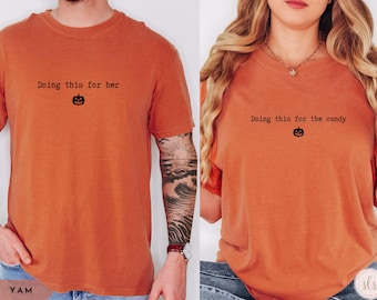 May include: Two orange t-shirts with the phrases "Doing this for her" and "Doing this for the candy" printed in black, each with a small pumpkin graphic. The shirts are a warm, autumnal color, perfect for Halloween or fall-themed events.