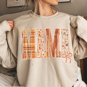 May include: Sand-colored sweatshirt with the word "MAMA" in large, decorative letters. The letters feature plaid, floral, and textured designs with bow accents. The sweatshirt has long sleeves.