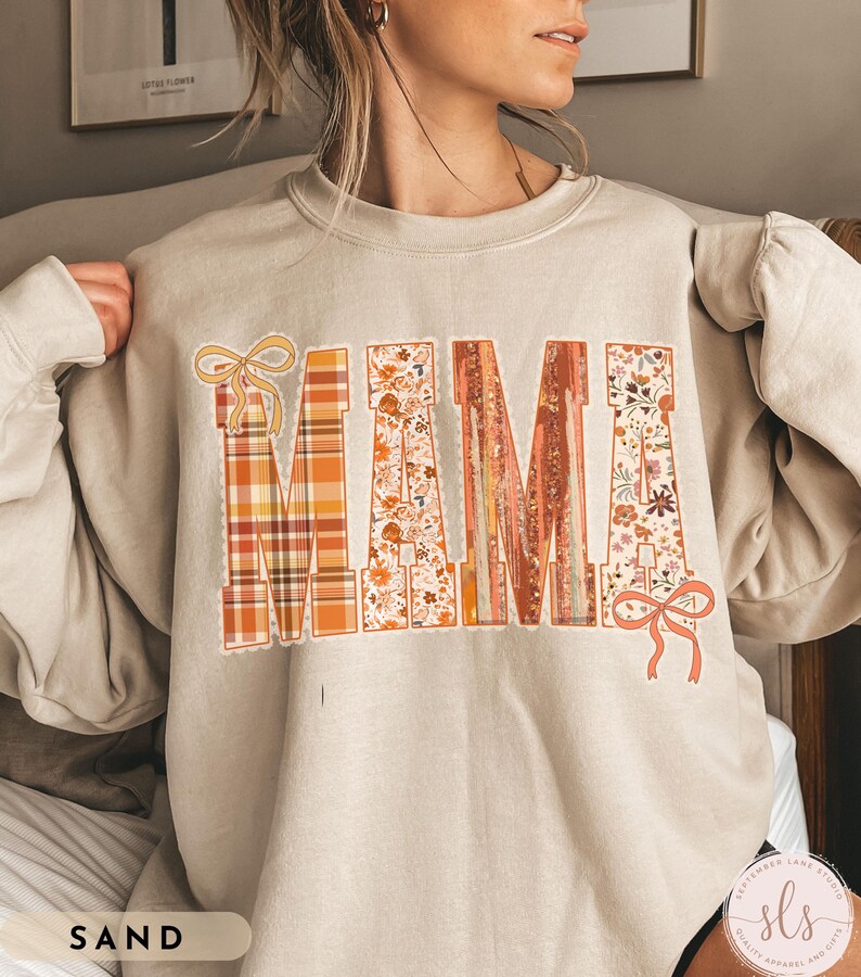 May include: Sand-colored sweatshirt with the word "MAMA" in large, decorative letters. The letters feature plaid, floral, and textured designs with bow accents. The sweatshirt has long sleeves.