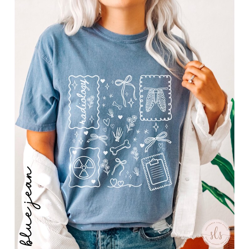 Puede incluir: Camiseta azul vaquero con un dise&ntilde;o gr&aacute;fico blanco que presenta la palabra "radiolog&iacute;a" e ilustraciones m&eacute;dicas. El dise&ntilde;o incluye huesos, corazones, lazos y un s&iacute;mbolo de radiaci&oacute;n.