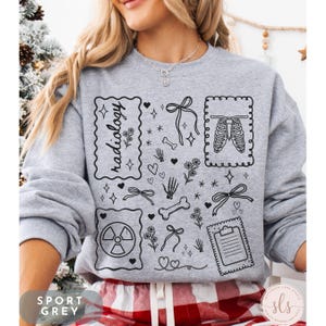Puede incluir: Sudadera gris deportiva con un dise&ntilde;o de radiolog&iacute;a en blanco y negro. El dise&ntilde;o incluye la palabra "radiolog&iacute;a", huesos, corazones, lazos, flores y una caja tor&aacute;cica. La sudadera tiene el texto "Sport Grey".