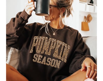 May include: Chocolate brown sweatshirt with the text "PUMPKIN SEASON" in a leopard print design. The sweatshirt is a relaxed fit and has long sleeves. A black mug is held up to the face. The bottom left corner of the image has the text "CHOCOLATE BROWN".
