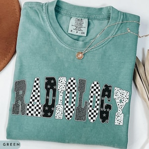 K&ouml;nnte beinhalten: Hellgr&uuml;nes T-Shirt mit dem Wort "RADIOLOGY" in einem auff&auml;lligen, gemusterten Design. Die Buchstaben zeigen eine Mischung aus karierten, Leopardenmuster- und Sternmustern. Eine goldene Halskette mit Anh&auml;nger liegt auf dem Shirt.
