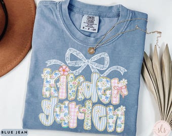 May include: A blue jean t-shirt with a floral design that says "Kinder Garden". The shirt has a bow and a small tag that says "Comfort Colors S-P-CH".