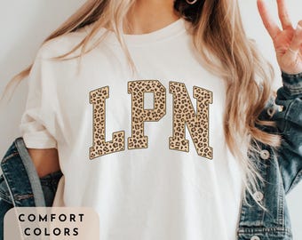 May include: A white t-shirt with the word "LPN" in brown leopard print letters. The shirt is a comfort colors shirt in ivory.