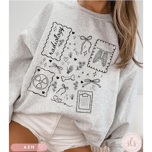 Puede incluir: Sudadera gris ceniza con ilustraciones en l&iacute;nea negra. El dise&ntilde;o incluye la palabra "radiolog&iacute;a", un s&iacute;mbolo de radiaci&oacute;n, huesos, lazos, flores y una caja tor&aacute;cica. La sudadera es de cuello redondo.