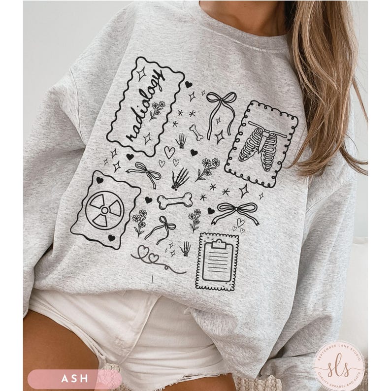 Puede incluir: Sudadera gris ceniza con ilustraciones en l&iacute;nea negra. El dise&ntilde;o incluye la palabra "radiolog&iacute;a", un s&iacute;mbolo de radiaci&oacute;n, huesos, lazos, flores y una caja tor&aacute;cica. La sudadera es de cuello redondo.