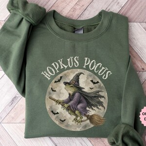 May include: A military green crewneck sweatshirt featuring a graphic of a frog dressed as a witch, wearing a pointed hat and purple cloak, riding a broomstick in front of a full moon with bats. The text "HOPKUS POCUS" is printed above the graphic.