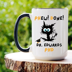 May include: White ceramic coffee mug with a black handle. The mug has a black and white illustration of a cat with the text "PHEW! DONE! DR. EDWARDS PHD" in orange.