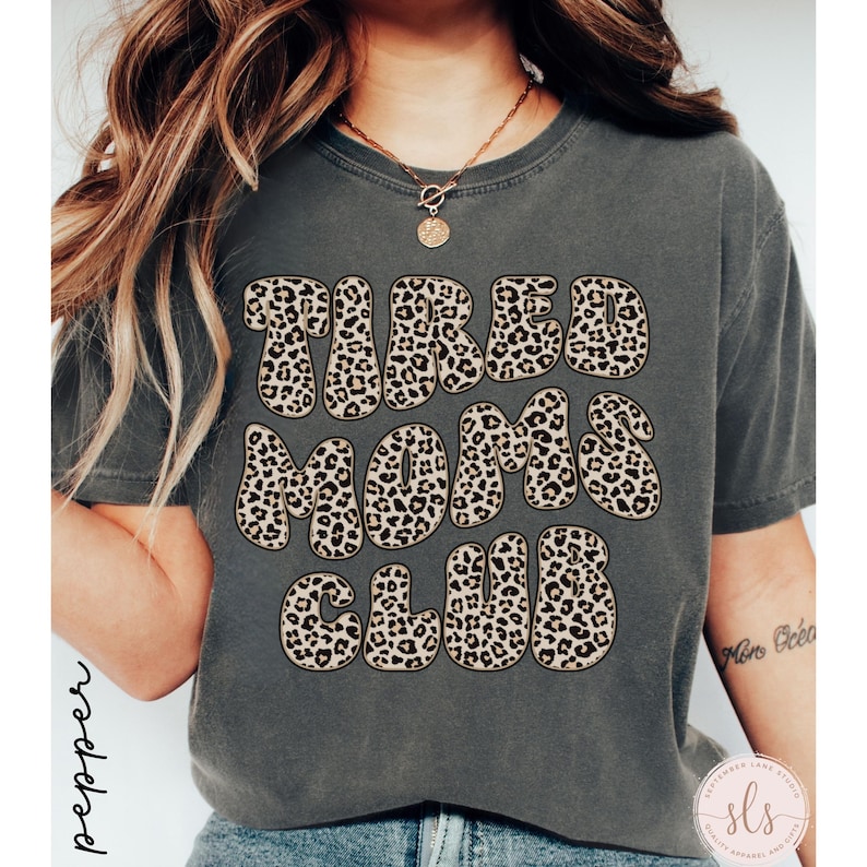 K&ouml;nnte beinhalten: Dunkelgraues T-Shirt mit dem Text "TIRED MOMS CLUB" in einem Leopardenmuster. Das Shirt hat kurze &Auml;rmel und einen Rundhalsausschnitt. Das Wort "pepper" ist vertikal auf der linken Seite aufgedruckt.